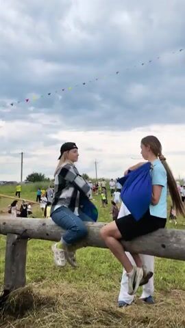 Two young women are sitting on a thick log that is elevated off the ground by supports. The woman on the left, wearing a black baseball cap, plaid shirt, and jeans, holds a blue fabric bag. The woman on the right, wearing a light blue t-shirt and black shorts, also holds a blue fabric bag. They are facing each other and appear to be engaged in some sort of game or competition, possibly a playful joust or tug-of-war with the fabric bags. Their legs are dangling over the sides of the log. Behind them, a crowd of people is gathered, and there are some wooden structures and flags in the background. The ground is covered with dry grass and hay. The sky is overcast.