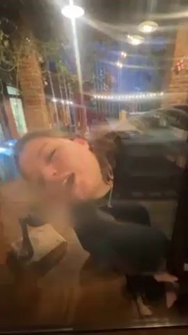 A woman with reddish hair is seen through a fogged-up window or glass surface in an indoor setting. The environment includes brick pillars, hanging lights, and what appears to be outdoor decorations, visible through the blurred glass. She speaks and gestures with her hand, as if interacting with someone on the other side of the glass.
