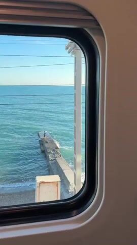 The video shows a view from a train window looking out at the ocean. The water is a bright turquoise color and there are small waves breaking on the shore. The beach is sandy and appears to be empty except for a few colorful beach towels. There is a concrete pier extending into the water on the left side of the frame, and a person is standing on it. A utility pole with wires is also visible near the shore. The sky is clear and blue. The train appears to be moving, as the scenery outside the window is subtly changing from frame to frame.