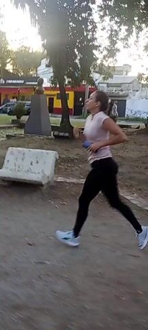 The video shows a woman jogging in an outdoor environment. She's wearing a light-colored t-shirt, black leggings, and light blue and white running shoes. She also appears to be carrying a phone or similar device in her hand. The setting looks like a park or a public area with trees and buildings visible in the background. The ground appears to be dirt or gravel.