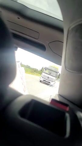 The video is shot from inside a vehicle, looking out through the rear window. A white semi-truck with a yellow tarp on its trailer is visible in the distance, driving on a road. The road appears to be in a rural or suburban area with trees and green vegetation visible on the sides. The sky is overcast. The interior of the vehicle is dark, with the back of seats and part of a speaker visible in the foreground. The lighting suggests it is daytime.