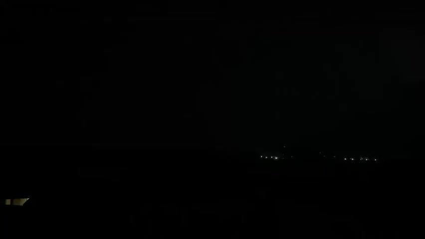The video captures a nighttime scene with a dark sky over a distant landscape. In the far distance, a faint line of lights suggests a settlement or urban area. A subtle, possibly distant, lightning strike illuminates the sky on the left side of the frame for a brief moment, casting a faint orange glow on the horizon. The foreground is mostly dark, with a portion of a building's window visible on the lower left corner. The overall impression is one of a storm approaching or passing by a remote location.
