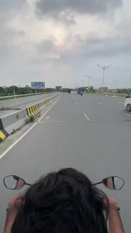 The video shows a first-person view from a motorcycle driving on a highway under a cloudy sky. The road is paved and has multiple lanes with white lane markings. Yellow and black striped barriers line the side of the road.  Other vehicles, including a van, a small truck, and an auto-rickshaw, can be seen driving in the distance. Street lights are spaced along the road. A billboard with "DPL" is visible on the left side of the road.  The back of the motorcycle driver's head and shoulders are visible, along with the motorcycle's rearview mirrors.