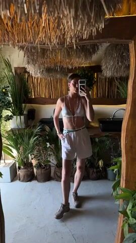 A woman is taking a mirror selfie in a brightly lit room with a tropical or bohemian theme. She is wearing a silver bikini top, white shorts, a green belt, and sneakers. She has her hair pulled back and is wearing necklaces and bracelets. The room is filled with plants and has a ceiling covered in straw-like material. There is a sink and faucet visible in the background, along with wooden elements and bamboo-like structures. The woman is posing and adjusting her hair.