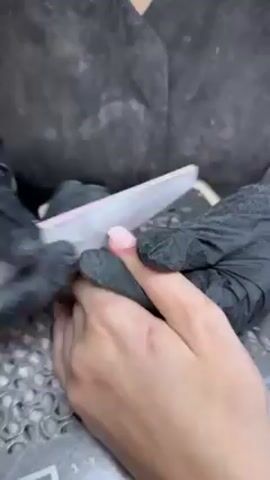 A person wearing black gloves is filing a person’s fingernail. The nail is pink. The manicurist is wearing a gray uniform. The manicure station appears to be messy with dust and shavings.