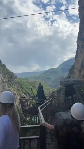 The video features a woman in a dark green dress, resembling a cloak, standing on a circular platform with a metal railing. She faces away from the camera, looking out over a mountainous landscape with lush green hills and a cloudy sky. Her arms are outstretched, as if gesturing or performing. In the foreground, two people wearing white hard hats are visible, with their backs to the camera, seemingly observing the woman. Another person with curly hair is partially visible in the bottom right, also looking towards the woman. The platform is connected to a path that leads into a dark cave-like structure on the right side of the frame. The overall scene appears to be an outdoor performance or photoshoot in a scenic natural environment.