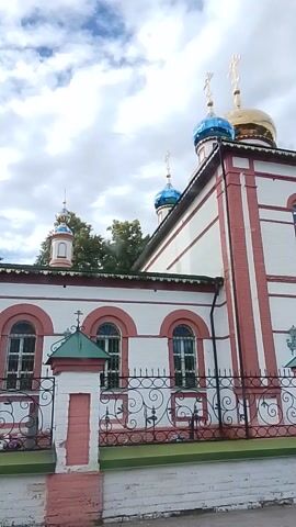 The video shows a side view of a church building with a bell tower. The building is predominantly white with pink trim. The bell tower is tall and slender, also with white and pink coloring, and features a green roof and a golden cross on top. Several onion domes are visible, some blue and some golden. The church has arched windows with wrought iron grills. There is a large, ornate black metal gate with golden angel decorations. The sky is cloudy. Trees are visible behind the church. The video is shaky and appears to be handheld.