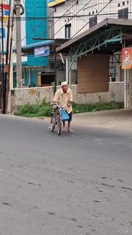 An elderly man with a white cap and light brown shirt is pushing a bicycle on a paved road. The bicycle is purple with a blue bag hanging from the handlebars. The man is barefoot and wearing dark shorts. He is looking to his right. In the background, there are buildings, a street light, and some greenery. The sky is visible above the buildings. The scene appears to be in an urban or semi-urban environment.