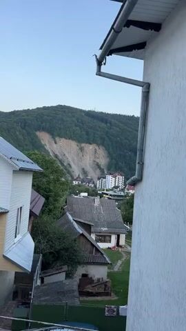 The video pans across a hillside covered in trees, with a large scar from a landslide visible on the upper portion. Below the landslide, a town with several buildings, including apartment complexes and smaller houses, is nestled into the trees. The foreground shows the roof of a house with a downspout, and the side of another house with white siding. The overall impression is a residential area in a mountainous, forested region, affected by a recent geological event.