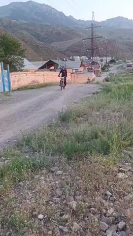 A person rides a dirt bike down a dusty, unpaved road. The road is lined with dry grass and rocks on either side. In the background, there are several buildings, some with corrugated metal roofs, and a tall electrical tower. The scene is set against a backdrop of rolling hills and mountains. The sky is clear and a light orange hue, suggesting it is either sunrise or sunset. The overall mood is peaceful and rural. There is no visible text in the media.