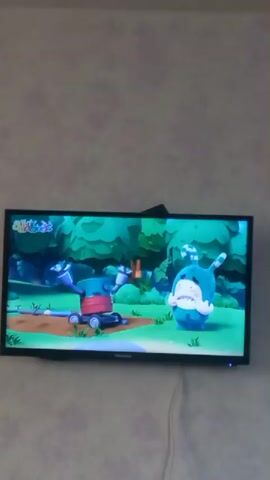 A cartoon character with blue fur and floppy ears is shown on a television screen. The character is sitting in a green, wooded area with trees and grass. The character is looking at a carrot on the ground. The scene is from a children's cartoon.