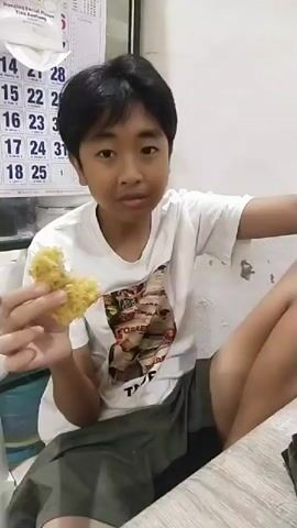 A young person wearing a white t-shirt with a graphic print on it and shorts is holding a phone and looking at the camera. The person is smiling and appears to be taking a selfie. A calendar is visible in the background.