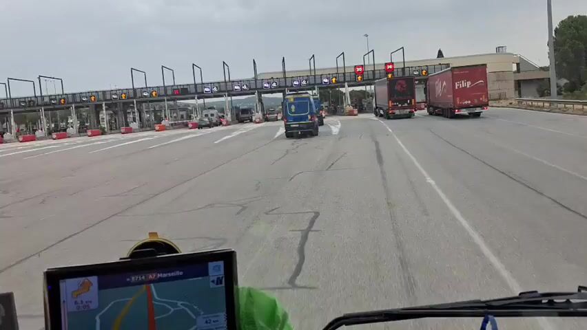 The video shows a first-person perspective from inside a vehicle approaching a toll plaza. The vehicle is in a lane with other vehicles, including several blue vans with yellow markings and two large red trucks. The toll plaza has multiple lanes, each equipped with overhead gantries displaying traffic signals and payment options. The sky is overcast, suggesting a cloudy day. The dashboard of the vehicle shows a GPS navigation screen displaying a map with a route highlighted. The vehicles are moving slowly, indicating a traffic queue at the toll booths.
