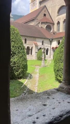 The video pans across a courtyard garden surrounded by historic stone buildings. The garden is rectangular with a gravel path running down the center, bordered by manicured green bushes on either side. In the middle of the garden, there is a small stone monument or fountain. The buildings appear to be part of a monastery or cathedral complex, with arched windows and doorways visible. One building has a large clock tower. The shot is taken from a covered walkway or cloister, with stone pillars in the foreground. The sky is partly cloudy.
