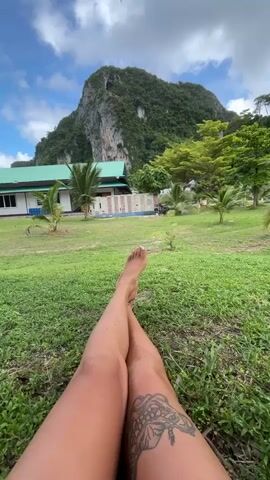 This is a first-person perspective video of a person lying on the grass with their legs crossed. The person's legs and feet are in the foreground of the image, showing a tattoo on the left thigh and an anklet on the right ankle. In the background, there is a grassy field, a building with a green roof, palm trees, and a large, rocky mountain. The sky is blue with some clouds.