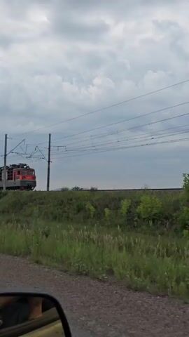 A red and gray electric train travels along a track through a grassy, overgrown field under a cloudy sky. The train has multiple cars, including a green hopper car. The view is from a car moving parallel to the train, with the side mirror visible in the foreground.