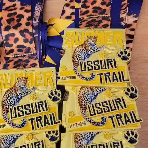 The video shows a close-up, overhead shot of a pile of yellow and blue "SUMMER USSURI TRAIL" race medals, each featuring a leopard illustration and paw prints. The medals are stacked in multiple rows and columns, with some lying flat and others slightly tilted. The lighting appears to be artificial, casting shadows on the medals. The surface beneath the medals is a wooden tabletop. A portion of a leopard-print fabric and a blue and yellow patterned ribbon are visible in the top left corner of the frame.