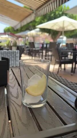 The video shows a close-up of a glass filled with ice and a slice of lemon on a wooden table. A hand is pouring Coca-Cola from a glass bottle into the glass. The beverage fizzes as it is poured, creating a light brown foam. The background is blurred but appears to be an outdoor seating area of a restaurant or cafe, with tables, chairs, and umbrellas visible. Sunlight filters through an awning, suggesting it is daytime. The scene is outdoors, likely in a warm climate, given the presence of umbrellas and greenery in the background. The overall mood is relaxed and refreshing, with the act of preparing a cool drink on a sunny day. No people are clearly visible, only the hand holding the bottle. The visible text on the bottle reads "Coca-Cola".