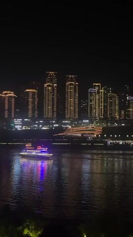 A nighttime scene unfolds along a river, with a city skyline illuminated by a multitude of lights in the background. Dominating the skyline are several tall, modern buildings, their facades adorned with rows of warm, golden lights, creating a striking visual against the dark night sky. Below these skyscrapers, a sprawling complex of buildings and structures stretches along the riverbank, also aglow with lights, suggesting a vibrant urban environment. In the foreground, the dark, rippling surface of the river reflects the colorful lights from the city and a prominent object on the water. A boat, brightly lit with red, white, and blue lights, cruises across the river, its illumination creating a vibrant dance of color on the water's surface. The lower portion of the frame shows a dark area, partially illuminated by faint green lights, suggesting foliage or the edge of the riverbank. The overall atmosphere is one of urban nighttime splendor and tranquility.