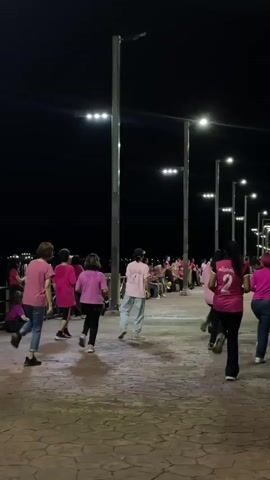 A group of women in pink shirts are dancing in a formation outdoors at night. They are on a paved walkway with lampposts providing light. The women are moving in unison, some facing the camera and others away from it. One woman in the foreground is wearing a pink shirt and blue jeans and appears to be leading the dance. Other women are wearing pink shirts and dark pants or skirts. The background is dark, with the lights from the lampposts creating a glow. There is a body of water in the distance. The overall atmosphere is festive and active.