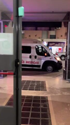 From behind a metal gate, a white ambulance with "URGENCIAS MEDICAS" in red letters on its side pulls up to the curb. The ambulance is marked with the number 29. Behind it, there is a white pickup truck parked by a building with a Milka chocolate sign. The sky is dark. There are square patterns on the ground.