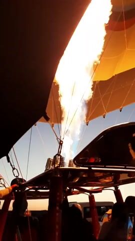 The video shows the inside of a hot air balloon basket. Flames are visible shooting from a burner, heating the balloon envelope above. The balloon is colorful with yellow, orange, red, and black stripes. The sky is visible in the background. Silhouettes of people can be seen in the basket, looking up at the balloon. The perspective is from within the basket, looking up towards the burner and the balloon.