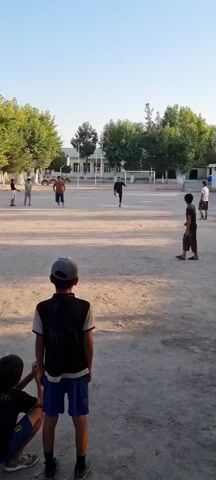 The video shows an outdoor scene on a dusty field, likely a schoolyard, in Havaskor, Tajikistan. The time of day appears to be late afternoon or early evening, with long shadows cast by the trees and buildings. The weather seems clear and warm, indicated by the bright sunlight and the light clothing worn by the people.

The central activity is a group of children playing soccer. Several children are scattered across the field, with one boy in the center actively kicking the ball. Others are observing or waiting for their turn. The background features a simple building, possibly a school, with trees lining the perimeter. There are also soccer goals visible, suggesting this is a designated play area.

In the foreground, two boys are positioned closer to the camera. One boy, wearing a black vest over a t-shirt and blue shorts, looks towards the camera with a slight smile. Another boy, partially visible, is sitting and looking away. The overall atmosphere seems relaxed and joyful, characteristic of children engaged in play.

There are no prominent signs or text visible in the video. The clothing of the children is casual, and they appear to be of various ages. The setting is rural or semi-rural, with unpaved ground and simple structures.