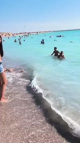 A woman in a black bikini top and denim shorts stands at the edge of the water on a crowded beach. She holds something in her hands and looks down at it. The water is a bright turquoise color, and many people are swimming and relaxing in it. In the distance, there are rows of umbrellas and beachgoers on the sand. The sky is clear and blue. The scene captures a typical sunny day at a popular beach.