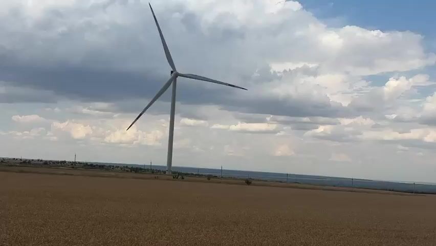 The video shows a field of wheat with several wind turbines in the background. The sky is partly cloudy, with a mix of blue sky and white clouds. The wind turbines are tall, white structures with three blades. They appear to be in operation, as the blades are rotating. The field of wheat is golden brown, indicating that it is ripe for harvest. The overall scene is one of a rural landscape, with a focus on renewable energy production.