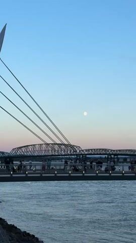 A wide shot captures a bridge stretching across a body of water under a twilight sky with a full moon visible. Several cables descend from the left, connecting to an elevated walkway, possibly part of a bridge or an observation deck, which is populated by a number of people. A boat, also filled with people, moves across the water from left to right. The water is choppy, and the sky transitions from a pale blue at the top to hues of pink and orange near the horizon. The bridge appears to be a truss bridge.