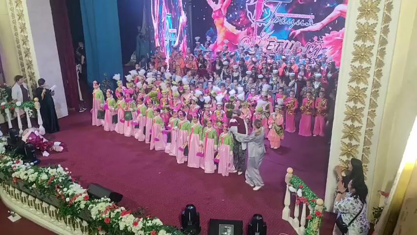 This media file captures a stage performance, likely a dance recital or cultural event, taking place in Qaskeleñ, Kazakhstan. A large group of children, dressed in colorful costumes, are lined up on a red carpeted stage in front of a vibrant LED screen displaying an abstract, energetic design with silhouettes of dancers and what appears to be Cyrillic text. Many of the children are wearing matching pink and green outfits, suggesting they are part of a dance troupe. Some adults, presumably instructors or organizers, are also present on the stage, interacting with the performers.

The setting is an indoor venue, evidenced by the stage lighting and the decorated backdrop. The audience area is visible in the foreground, with some people observing the event. The overall atmosphere appears celebratory and organized, with the focus on the young performers. The visual style of the LED screen and the ornate golden trim on the right side of the frame suggest a formal or festive occasion. The visible text on the screen is in Cyrillic, which is consistent with the location of Kazakhstan. The lighting suggests the event is taking place during the evening or in a dimly lit hall. The emotions conveyed are primarily those of anticipation and excitement, common at such performances.