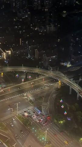 The video shows an aerial view of a city at night. The foreground features a multi-level intersection with cars driving on the roads. Traffic lights are visible, along with streetlights illuminating the area. Elevated roadways curve through the scene, with cars traveling on them. In the background, numerous buildings are lit up, creating a sprawling urban landscape. The overall impression is one of a busy, illuminated city at night.