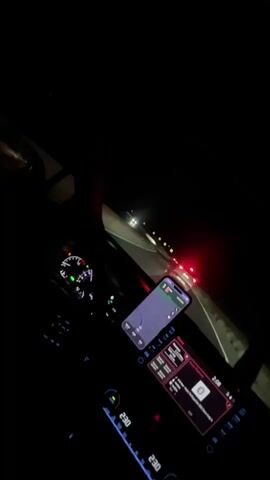 This is a first-person perspective video taken at night from inside a car. The driver's dashboard is visible, with illuminated gauges and a navigation system on a smartphone mounted to the center console. The car's interior lights are also on. The view through the windshield shows a dark, winding road with other cars visible in the distance, their headlights and taillights creating streaks of light. Yellow road signs are also visible along the side of the road. The overall impression is of driving at night, possibly on a highway or rural road.