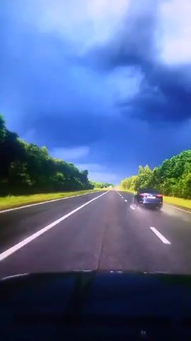 This video captures a forward-facing view from a vehicle driving on a highway. The road is paved with white lines marking the lanes. On either side of the road, there are dense green trees forming a natural barrier. The sky above is a dramatic, dark blue with heavy, ominous clouds, suggesting an impending storm. In the distance, a white truck is visible driving on the highway. The overall scene conveys a sense of movement and anticipation, with the approaching weather creating a striking backdrop.