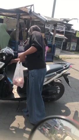 The video shows a street scene with a food stall set up on the back of a truck. A man is bent over behind the counter, seemingly preparing food. Two women and a young girl are standing in front of the stall, interacting with the man. The girl is wearing a pink dress. A motorbike is parked in the foreground, obscuring part of the scene. The environment appears to be outdoors, possibly a market or roadside.