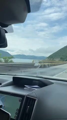 The video offers a first-person view from the dashboard of a car driving along a scenic road. Inside the vehicle, parts of the black dashboard are visible, along with an infotainment screen displaying "CarPlay," two temperature readings of "22.0°C" and "22°C," and the time "15:42." A lower screen shows a navigation map. A rearview mirror hangs from the top of the windshield.

The exterior view shows a multi-lane highway, possibly a bridge or causeway, running alongside a large body of water, likely the Danube River in the vicinity of Drobeta-Turnu Severin, Romania, given the location context. On the right side of the road, green, forested mountains rise in the distance. The road is bordered by concrete barriers topped with yellow and black railings that separate it from the water. Another vehicle, possibly a truck, can be seen in the distance traveling in the same direction. The sky is partly cloudy with visible patches of blue, indicating a bright daytime setting. The overall scene depicts a pleasant drive through a mountainous and watery landscape.
