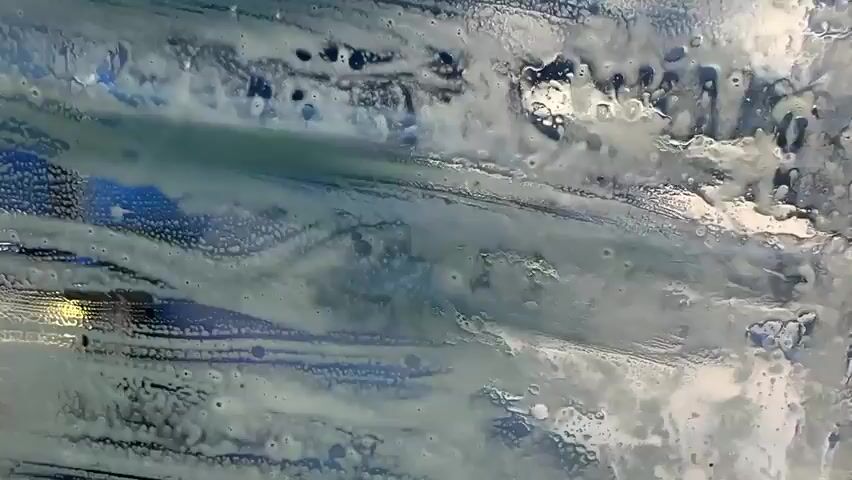 The video shows a car's windshield from the inside, covered in soap suds and water, as it moves through an automated car wash. The suds are white and foamy, streaking across the glass in various patterns, with some areas showing blue and black underneath. The car appears to be moving forward, with the car wash machinery visible as blurred shapes outside the vehicle. The interior of the car is dark in the foreground, with parts of the dashboard and windshield frame visible. The overall impression is one of being inside a car during a wash cycle.