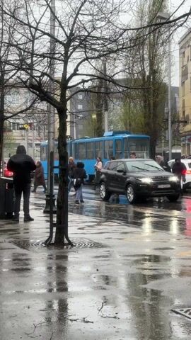 The video captures an urban street scene on a rainy day. The ground is wet, reflecting the light, and people are seen crossing a street, some holding umbrellas.

In the foreground on the left, a bare tree trunk stands, partially obscuring the view. The street itself is wet, with reflections visible on its surface.

Throughout the video, various pedestrians are crossing the street from different directions.
- At the beginning, a black SUV, possibly a Range Rover, with its headlights on, is driving from right to left across the crosswalk. Its license plate, "ZG 413-VZ", is partially visible.
- Behind the SUV, a blue tram is visible in the background, either stationary or moving slowly.
- Several people are crossing the street. One woman, prominent in the mid-section of the video, is wearing a grey coat and a light-colored scarf, possibly a hijab, and holding a red umbrella. She appears to be smiling slightly.
- Other pedestrians are dressed in dark, practical clothing, including hooded jackets and coats, suitable for rainy weather. Some are carrying bags or umbrellas.
- Towards the end of the clip, a man in a dark brown jacket and light grey pants walks into the frame from the right.

The overall atmosphere is that of a typical, busy city street during overcast, rainy daytime conditions. Buildings are faintly visible in the blurry background, further emphasizing the urban setting. The video focuses on the movement of people and vehicles through the wet street environment.