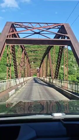 The video captures a first-person perspective from inside a vehicle driving across an old, reddish-brown metal truss bridge. The bridge features a prominent overhead and side lattice-work of steel beams, forming a tunnel-like structure over the roadway. The road surface appears to be asphalt, flanked by low concrete barriers and metal railings. Lush green, tree-covered mountains or hills rise on either side of the bridge, indicating a mountainous or hilly landscape. A clear blue sky is visible through the bridge's superstructure. Bright sunlight casts distinct shadows from the bridge's framework onto the road. The dashboard and the front edge of the vehicle's hood are visible at the bottom of the frame. No people or text are visible. The scene takes place during a sunny day in Bzypta, Abkhazia.