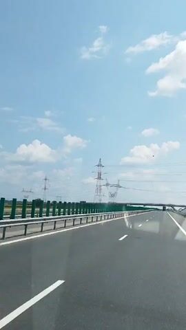 A white car is seen from the perspective of another vehicle driving on a highway. The highway has multiple lanes and is bordered by a guardrail and green fencing. Tall power line towers are visible in the background, along with a bridge crossing over the highway. The sky is blue with scattered white clouds. Another white vehicle, possibly a van, is ahead of the white car.