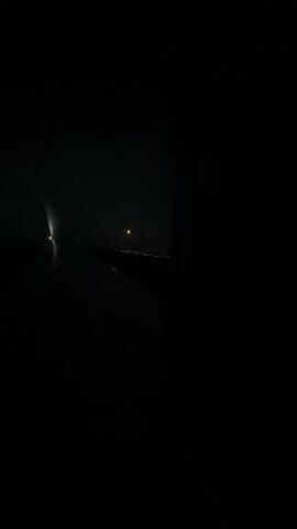 The video shows a view from inside an airplane at night. Through the window, a blurry orange moon is visible in the dark sky. Below the moon, indistinct city lights glimmer in the distance. The airplane's wing is partially visible in the foreground. The scene is dark and slightly out of focus.