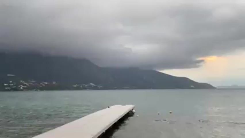 The scene captures a cloudy day by the sea.  A long, white pier extends into the water, with a backdrop of mountains shrouded in low-lying clouds. The sky is overcast with hints of sunlight peeking through towards the right side.  The water is calm and slightly choppy, with a few yellow buoys visible near the pier.  The mountains are covered with some greenery, punctuated by houses and buildings along the coastline.