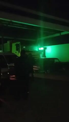 The video shows a parking lot or staging area under a bridge at night. There are several vehicles parked, including at least two vans, a car, and a truck, possibly a semi-truck.  Red lights are prominently visible, likely from the truck. The area is poorly lit overall, with some green and white light sources adding to the illumination. There is at least one person, a dark silhouette, walking around the vehicles. The focus is somewhat blurry and unstable, particularly at the beginning of the video.
