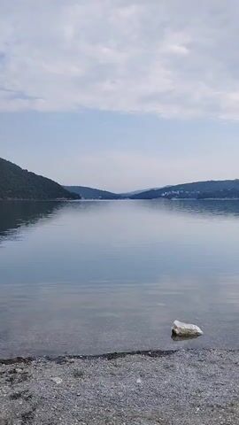 The video shows a serene lake with a slight ripple on the surface. The water is calm and reflects the sky and the surrounding hills. The shoreline is made of small pebbles. A single rock sits on the shore near the water's edge. The sky is partly cloudy, with soft light filtering through. Hills covered in green foliage rise from the opposite side of the lake, disappearing into the distance. There are no visible people or animals in the frame. The overall impression is one of tranquility and natural beauty.
