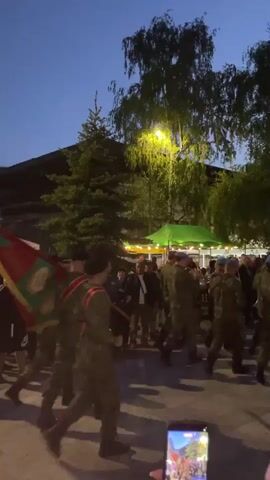 The video shows a group of people in military uniforms marching through a public square. They are carrying a flag with a red and green design. People are standing around watching the procession. The event is taking place in the evening, as the sky is a dusky blue. The area is paved and lined with trees and buildings. A phone recording the event is visible in the foreground.