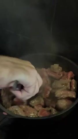 A person's hand is stirring diced meat in a frying pan. The meat is mostly brown with some pink in the center. Steam is rising from the pan, indicating it is hot. The pan is on a black stovetop.