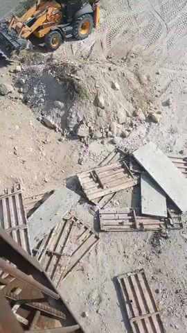 The video begins with a close-up of a rusty metal bar and a crane hook with a red component. The camera then pans upwards, revealing a construction site. In the background, there is a yellow backhoe loader parked on a dirt embankment. The foreground shows scattered wooden pallets and debris. As the camera continues to pan and zoom out, the construction site becomes more visible. There are piles of dirt and rocks, more wooden pallets, and sections of concrete. Further in the background, a large corrugated metal building with scaffolding is under construction. There are also various industrial machines, including a large yellow cylindrical machine, a smaller machine with blue components, and what appears to be a water tower. The scene depicts an active construction or demolition area.