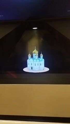 The video shows a holographic projection of a blue and gold domed church. The projection is presented within a dark, pyramid-shaped enclosure with a light source illuminating the top. The church appears to be rotating slowly on a white circular base. The lower portion of the image shows a beige surface, likely part of the display enclosure.