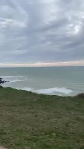 The video shows a view of the ocean from a grassy clifftop under a cloudy sky. The sea is choppy with small waves visible along the shoreline. A few birds fly overhead. There's a low fence and some shrubs along the edge of the cliff. In the far right of the video, you can see a portion of a concrete structure. The camera pans slowly.