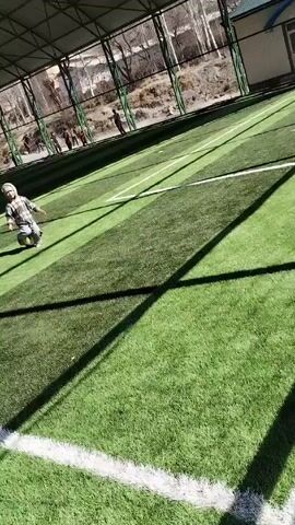 A child wearing a hooded jacket, jeans, and sneakers is shown tripping and falling while attempting to kick a blue and yellow soccer ball on a green turf field. The child slides a short distance. The field is enclosed by a fence and a metal structure that casts shadows on the field.  A white line marking the boundary of the field is visible. In the background, there are several other people standing or walking. The video is shot from an elevated position.
